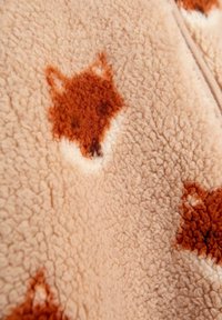 Švelnus smėlio spalvos fleece audinys su oranžiniais, išsibarsčiusiais lapės formos raštais. Paviršius atrodo šiltas ir jaukus.