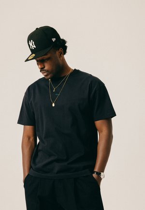 Mann trägt eine schwarze New York Yankees-Kappe, ein schwarzes T-Shirt, goldene Halsketten, eine Uhr und schwarze Hosen, schaut nach unten und hat die Hände in den Taschen.