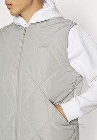 Gesteppte, hellgraue Weste mit hohem Kragen und durchgehendem Reißverschluss. Darunter eine weiße Kapuzen-Sweatshirt, das eine sichtbare Tasche zeigt.