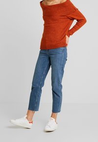 Pull en maille orange à épaules dénudées avec un col côtelé, associé à un jean bleu cropped et des baskets blanches avec des accents rouges.