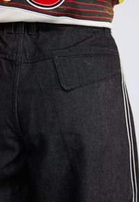 Calças de ganga pretas com um bolso traseiro, cintura plana e riscas brancas laterais. O tecido possui uma textura suave com um brilho subtil.