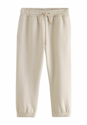 Ragazzo in piedi con le mani in tasca, indossa una maglietta bianca, pantaloni beige chiaro e sneakers bianche con logo Nike marrone davanti a un muro beige.