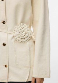 Kreemikas jakk suure esitasku, pruunide nuppude ja paremal vöökohal oleva kangasroosiga.