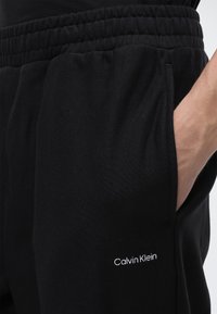 Κοντινό πλάνο ενός ατόμου που φοράει μαύρη φόρμα Calvin Klein με ελαστική μέση και το χέρι στην πλαϊνή τσέπη.