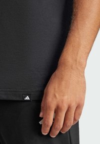 Μαύρο βαμβακερό T-shirt με εφαρμοστή γραμμή και κοντά μανίκια, διαθέτει λευκή ετικέτα λογότυπου Adidas στη κάτω άκρη. Χέρι μερικώς ορατό.