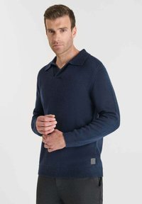 Suéter de color azul marino, de manga larga, con cuello tipo polo y puños acanalados. Textura suave con un parche de logo cerca del dobladillo. Diseño de corte recto.