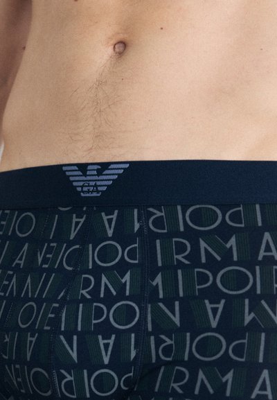 Muške bokserice s tamnim pojasom na kojem se nalazi logo. Tamni uzorak s tekstom u zelenim i sivoj boji, glatka tekstura tkanine.