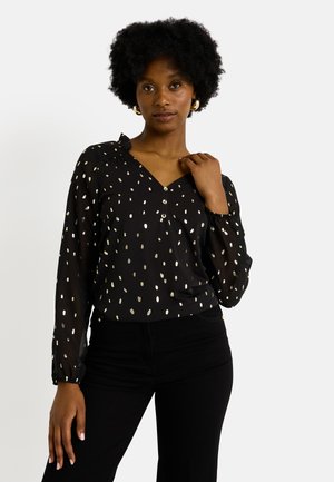 Blouse noire avec motif de pois argentés, décolleté en V, manches longues avec poignets élastiques et détails de boutons à l'avant. Texture de tissu lisse.