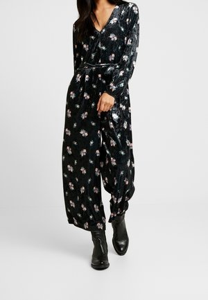 Schwarzer Samt-Overall mit langen Ärmeln, der mit einem floralen Muster in Pink und Grün verziert ist, tailliert und mit lockerer Passform, kombiniert mit schwarzen knöchelhohen Stiefeln.