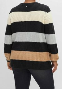 Persona che indossa un maglione lavorato a maglia spesso con larghe righe orizzontali in nero, beige, bianco e grigio, abbinato a jeans scuri, voltata di spalle.
