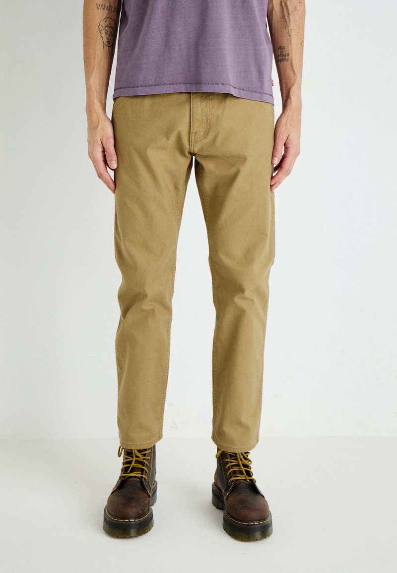 Pantalones de algodón color caqui claro con un ajuste regular, combinados con botas de cuero marrón oscuro con cordones amarillos y una suela gruesa.