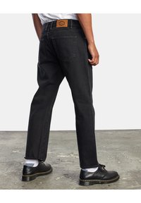 Vaqueros de mezclilla negros de corte recto, con costuras visibles y una etiqueta de cuero en la cintura. Combinados con zapatos negros y calcetines blancos.