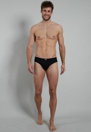 Black swim briefs made of stretchy fabric, featuring a low-rise design and a logo on the waistband, worn by a standing male model.