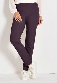 Vrouw in een strakke donkerpaarse broek en witte sneakers, geschilderd met gekruiste benen tegen een effen lichte achtergrond.