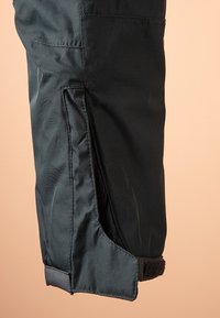 Gros plan d'une jambe de pantalon noire avec sangle Velcro ajustable et coutures renforcées sur fond beige.