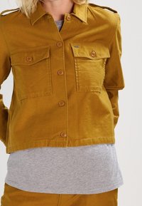 Senapsgul knäppt jacka med två bröstfickor, axelremmar och en kortad design, bärs över en grå t-shirt.