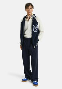 Marineblau und weiße Fleecejacke mit geripptem Kragen und großem 'G'-Emblem, kombiniert mit einem weißen Poloshirt, marineblauen Hosen und blauen Turnschuhen.