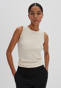 Haut sans manches beige à côtes avec un col rond, design ajusté et texture lisse, associé à un pantalon noir à coupe décontractée.
