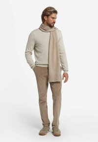 Un pull en laine beige associé à un pantalon en velours côtelé marron clair. Une longue écharpe douce dans une teinte similaire est négligemment drapée autour du cou.