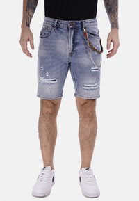 Pantalones cortos de mezclilla azul claro con detalles desgastados, bordes deshilachados y un accesorio de cadena adjunto. Combinados con zapatillas blancas.