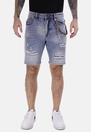Pantalones cortos de mezclilla azul claro con detalles desgastados, bordes deshilachados y un accesorio de cadena adjunto. Combinados con zapatillas blancas.
