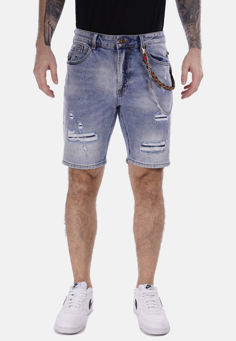 Pantalones cortos de mezclilla azul claro con detalles desgastados, bordes deshilachados y un accesorio de cadena adjunto. Combinados con zapatillas blancas.