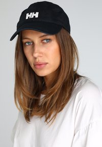 Zwarte baseballpet met geborduurd wit "HH" logo op de voorkant. Zacht materiaal, gebogen klep en verstelbare band aan de achterkant.