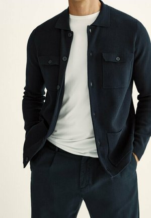 Mand iført en navyblå cardigan med knapper over en hvid t-shirt og matchende navy bukser, hænderne i lommerne, foran en neutral baggrund.