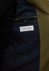 Interno della giacca verde oliva con una fodera in raso blu scuro. Presenta una tasca con un'etichetta bianca che riporta la scritta "PIER.ONE" e una chiusura a bottone.