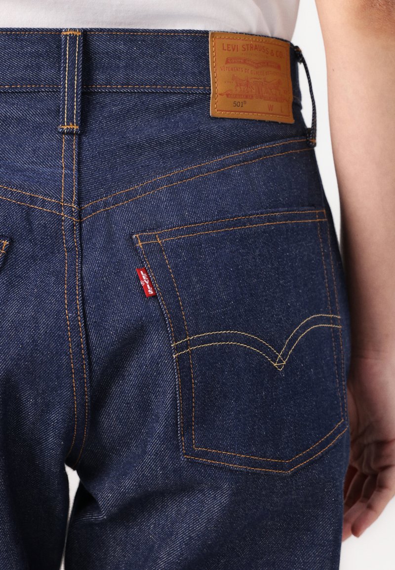 Granatowe dżinsy z ciemnego denimu z skórzaną metką w pasie, pomarańczowymi szwami i charakterystycznymi tylnymi kieszeniami z czerwoną metką Levi's.