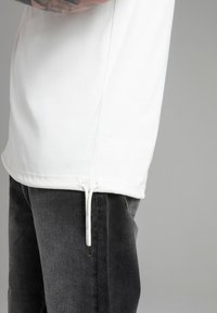 T-shirt bianca a maniche corte con orlo regolabile, indossata sopra jeans grigio scuro. Tessuto morbido con una vestibilità rilassata.