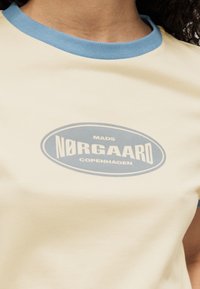 Μπλούζα από βαμβάκι σε κρεμ χρώμα, με ανοιχτό μπλε ριμπ λαιμόκοψη και γραφικό τύπωμα σε μπλε και λευκό, με το κείμενο "MADS NØRGAARD COPENHAGEN".