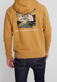 Sudadera amarilla para hombre con un logo de camuflaje en la espalda y el texto "NUNCA DEJES DE EXPLORAR". Presenta un ajuste cómodo y puños acanalados.