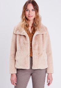 Veste en fausse fourrure beige avec un col classique, des poches latérales et une texture douce. Associée à une chemise marron à boutons et à un pantalon à carreaux.