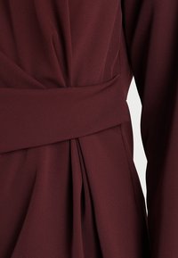 Tessuto di colore bordeaux con una texture liscia, caratterizzato da una cintura larga e pieghe verticali, che evidenziano un design strutturato.