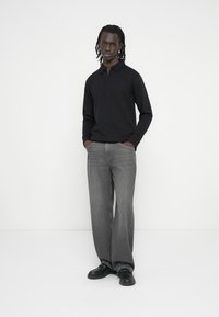 Μαύρο πόλο με μακριά μανίκια και γιακά με φερμουάρ, συνδυασμένο με ξεβαμμένα γκρι τζιν φαρδιάς γραμμής και μαύρα παπούτσια, σε απλό λευκό φόντο.