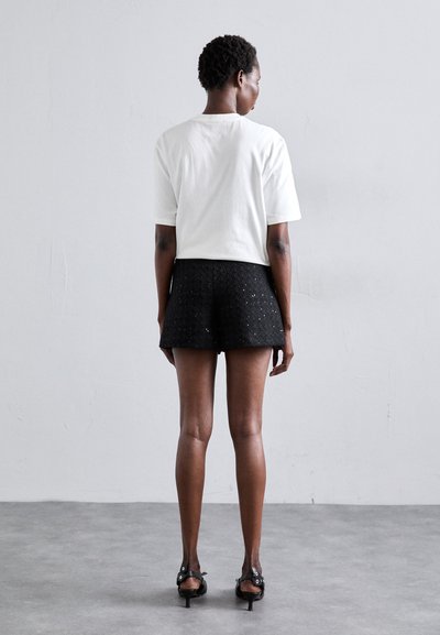T-shirt en coton blanc, à manches courtes, vue de dos ; associé à un short noir texturé avec un motif géométrique, complété par des sandales à talons.