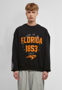 Fekete, oversized kapucnis pulóver narancssárga "Florida University 1853" felirattal és grafikai elemekkel, bordás mandzsettával és aljjal.