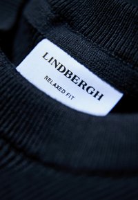 Schwarzer gerippter Stoff mit einer Nahaufnahme eines Kleidungslabels, auf dem "LINDBERGH" und "RELAXED FIT" in weißem und goldenem Text steht.