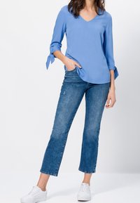 Blouse bleu clair à col en V avec des manches 3/4 et des détails à nouer, associée à un jean bleu ajusté et des baskets blanches.