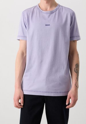 Personne portant un t-shirt à manches courtes violet clair avec le texte "BOSS" sur la poitrine et un pantalon noir sur un fond uni.