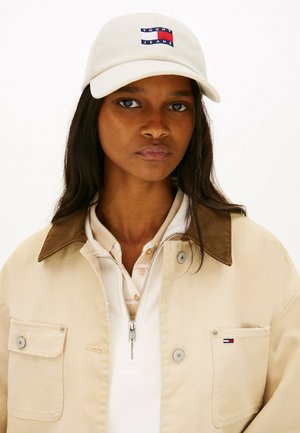Jeune femme aux cheveux foncés portant une veste beige avec un col marron et une casquette blanche arborant le logo Tommy Jeans à l'avant.