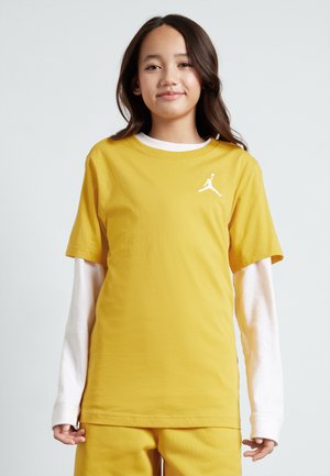 Camiseta de algodón amarilla con cuello redondo y mangas cortas, que presenta un logotipo blanco de Jordan en el lado izquierdo del pecho. Llevada sobre una camisa blanca de manga larga.