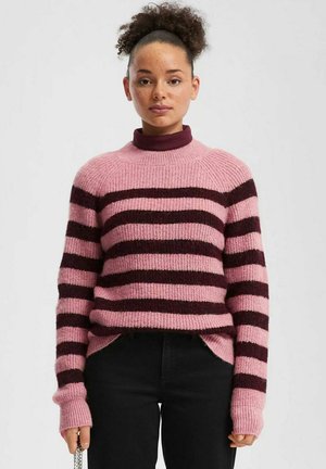 Różowy sweter w paski ciemnego burgundowego koloru, z wysokim kołnierzem, prążkowaną teksturą, długimi rękawami i luźnym krojem. Noszony z czarnymi spodniami.