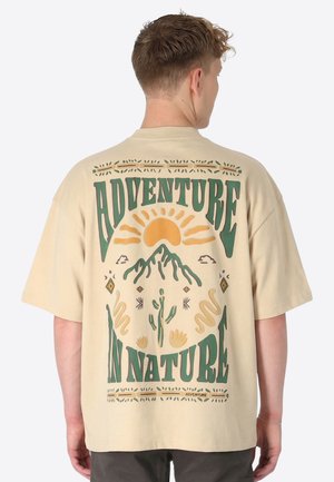 Mann trägt ein beiges T-Shirt mit grünem und orangefarbenem Motiv auf dem Rücken, das Sonne, Berg, Kaktus und den Schriftzug "Abenteuer in der Natur" zeigt.