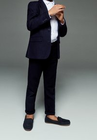 Giacca da completo blu navy sopra camicia bianca, abbinata a pantaloni scuri e mocassini. Tessuto liscio con una vestibilità sartoriale, caratterizzata da bottoni decorativi.