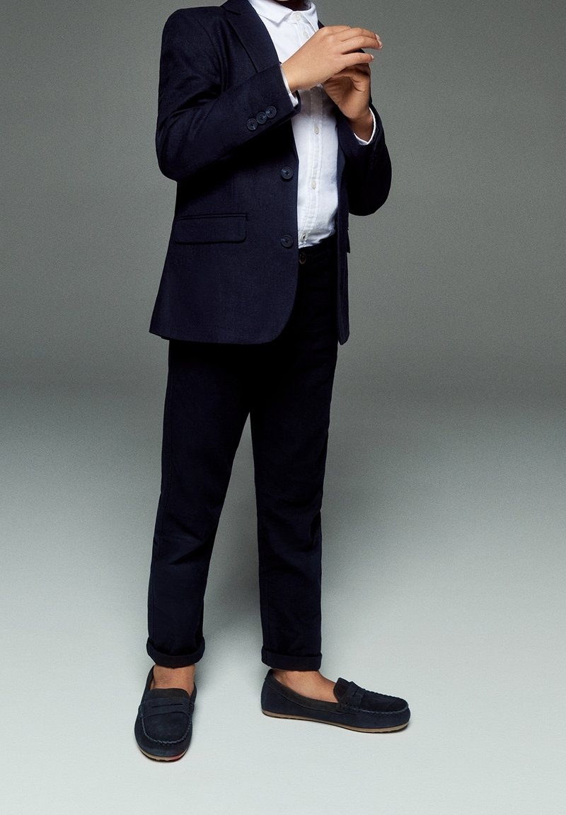 Giacca da completo blu navy sopra camicia bianca, abbinata a pantaloni scuri e mocassini. Tessuto liscio con una vestibilità sartoriale, caratterizzata da bottoni decorativi.