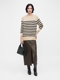 Femme aux cheveux courts et foncés portant un pull rayé beige et noir, une jupe en cuir marron avec une fente à l'avant, des bottes noires, tenant un sac à main noir.