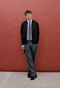 Veste bomber noire sur une chemise à rayures claires, associée à un pantalon gris plissé et des chaussures en cuir noires. Se tenant devant un fond rouge.
