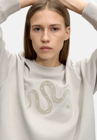 Jasnokremowy sweatshirt z fakturowanym wzorem węża na przodzie, z pluszowymi akcentami i ściągaczami. Gładki materiał o luźnym kroju.